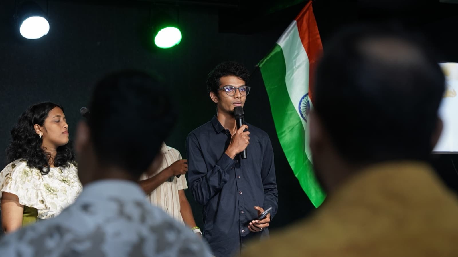 A young man speaking into a microphone beside an Indian flag